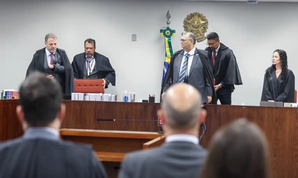 Homens em togas e ternos em um ambiente de tribunal, com a bandeira do Brasil e o brasão nacional ao fundo.