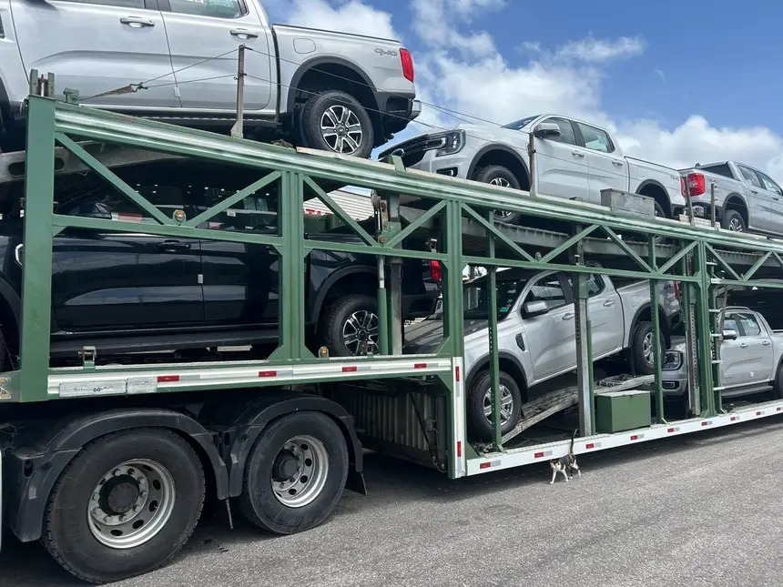 Caminhão cegonha transportando várias picapes Ford Ranger de cores prata e preta sob céu azul com nuvens.