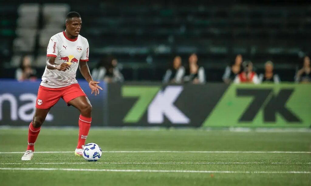 Jogador de futebol em campo com uniforme branco e vermelho, controlando a bola.