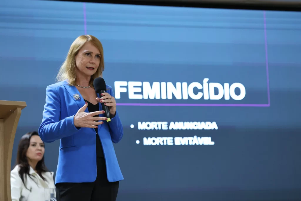 Mulher em blazer azul falando ao microfone com "FEMINICÍDIO" e pontos-chave no fundo.