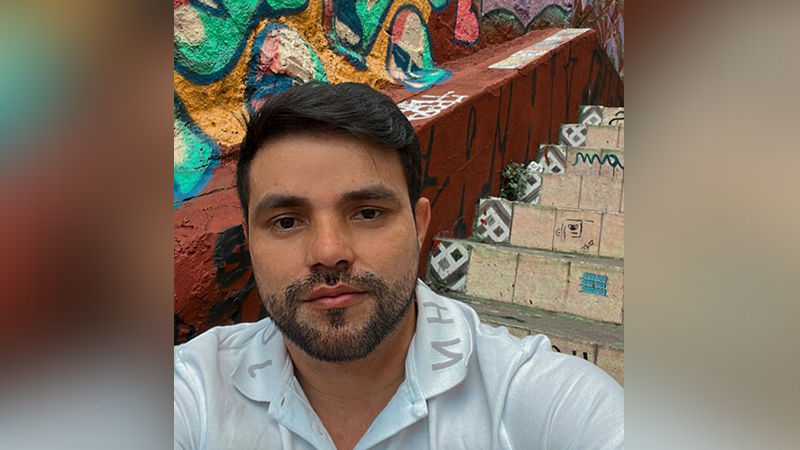 Homem com barba posando em frente a muro grafitado e escadaria artística.