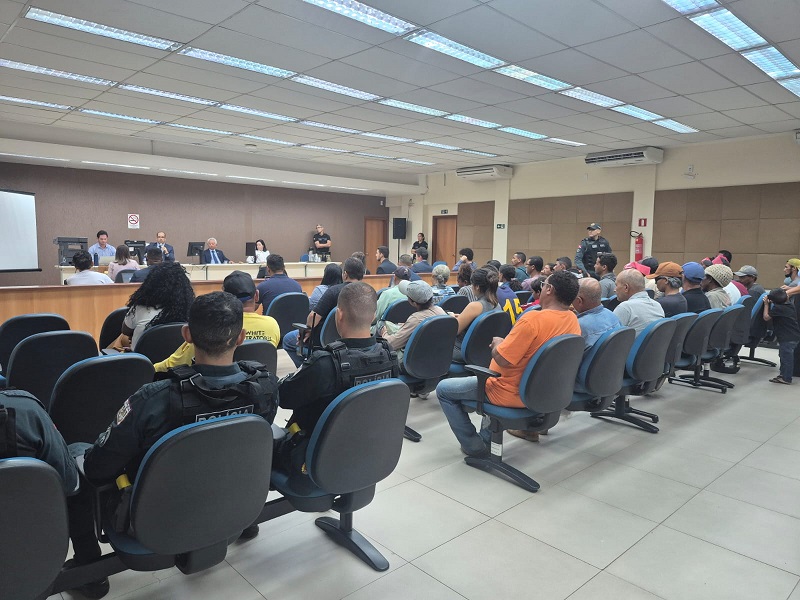 Pessoas assistindo a uma audiência ou reunião em um salão com oficiais de segurança.