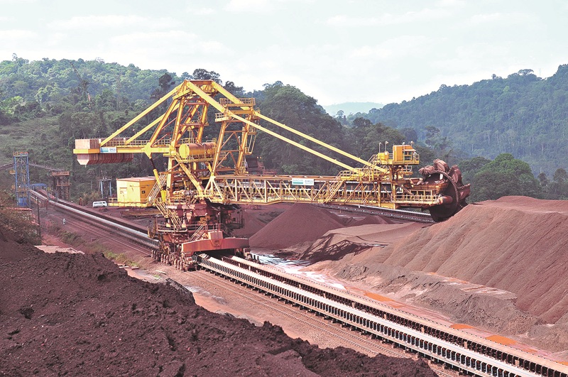 Máquina de mineração industrial amarela em operação com grandes pilhas de minério e floresta ao fundo.