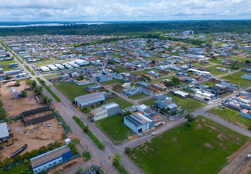 Vista aérea de uma cidade com casas, lotes vazios e floresta ao fundo, com um rio.