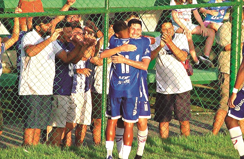Dois jogadores de futebol se abraçando em campo com torcedores aplaudindo ao fundo.