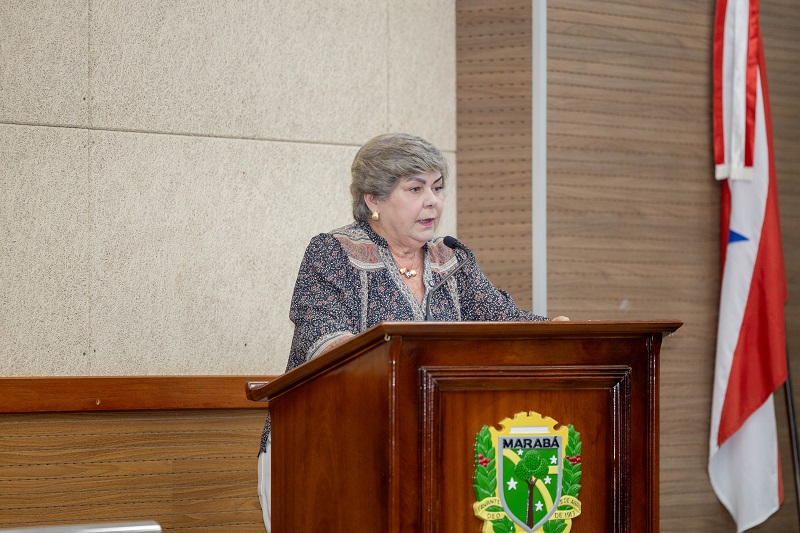 Mulher de cabelos grisalhos falando em púlpito com brasão de Marabá.