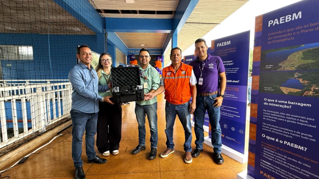 Grupo de cinco pessoas, incluindo dois homens e uma mulher entregando uma maleta de equipamentos, com painel sobre PAEBM ao fundo.