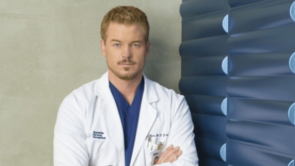 Ator Eric Dane em um jaleco médico e scrubs azuis, olhando para frente.