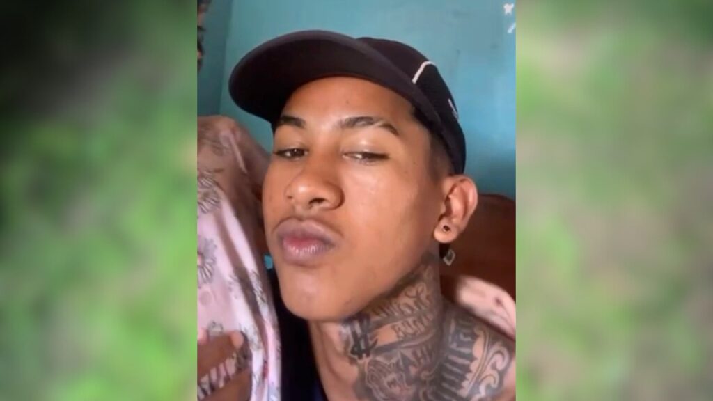 Jovem de boné preto e muitas tatuagens no pescoço olhando para a câmera.