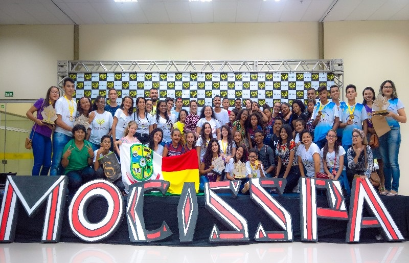 Grupo grande de estudantes e adultos celebrando em evento, com letras 'MOLISSFA' e alguns troféus.