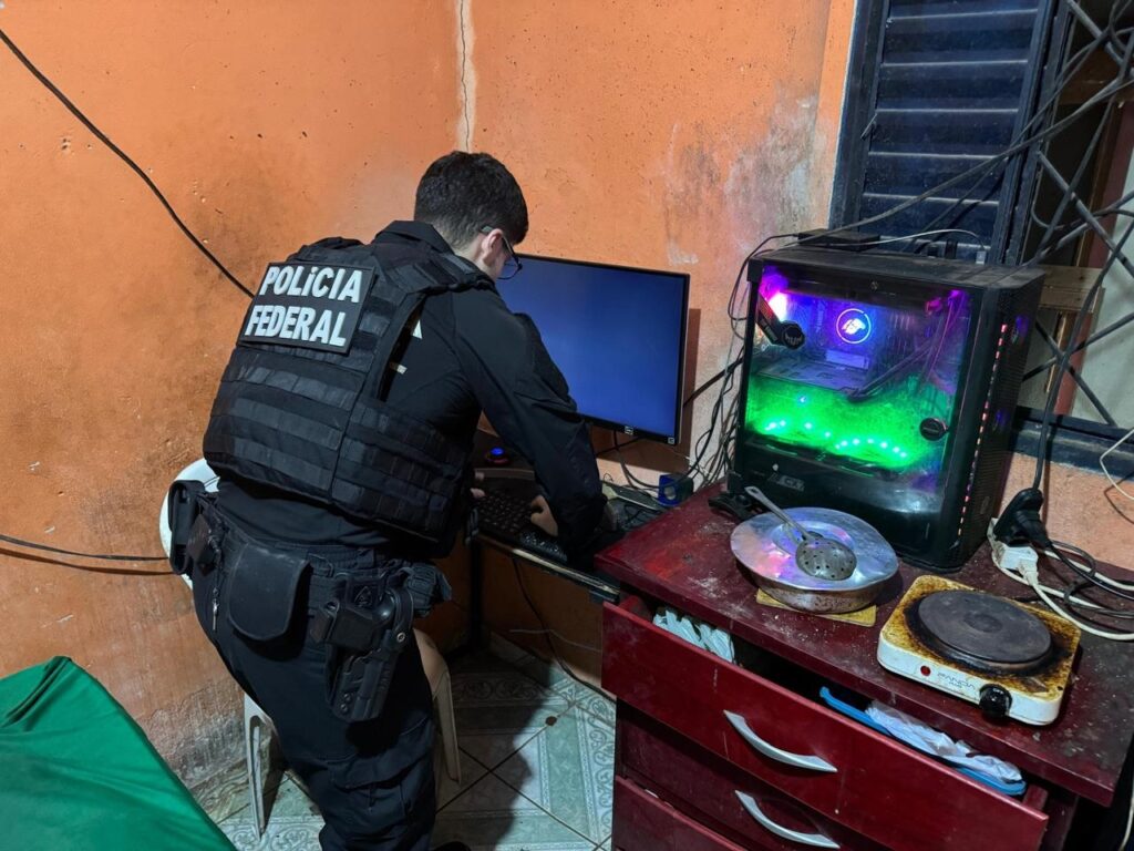 Um policial federal de costas, vestindo colete tático, opera um computador durante uma investigação.