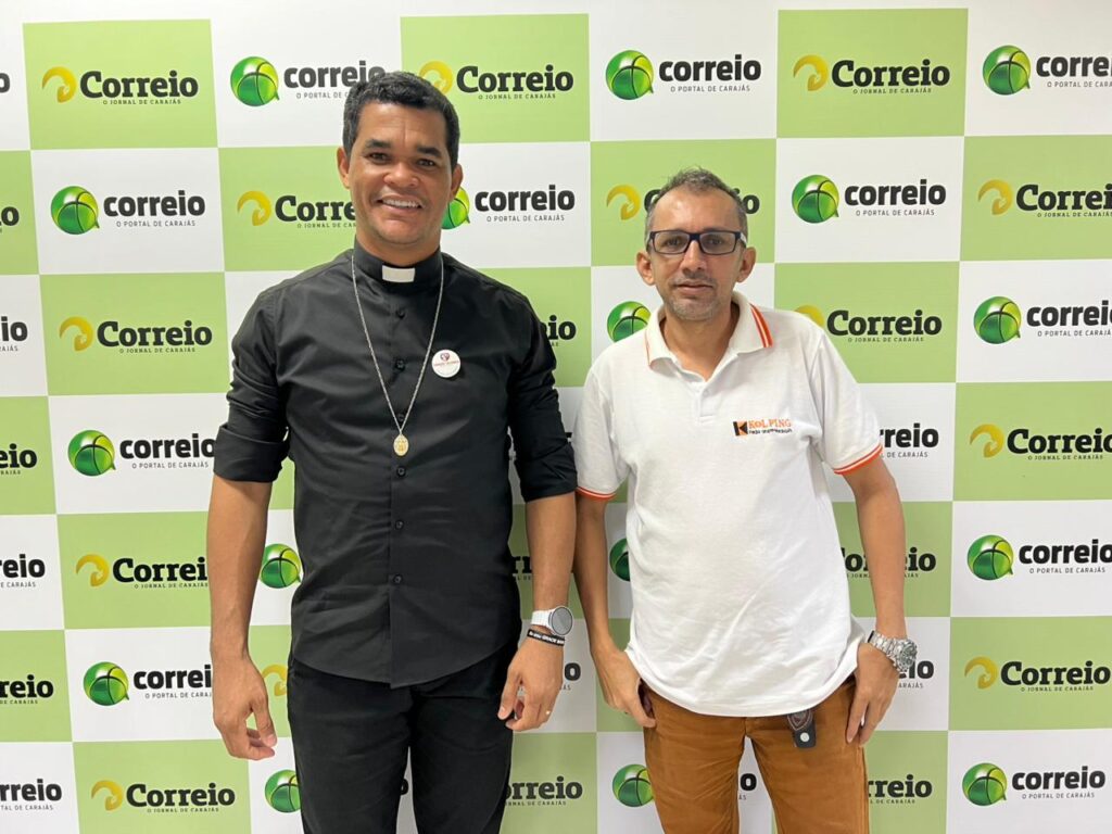 Dois homens, um padre e outro de camisa polo, sorrindo em frente a um fundo de parede com o logo 'Correio' em verde e branco.