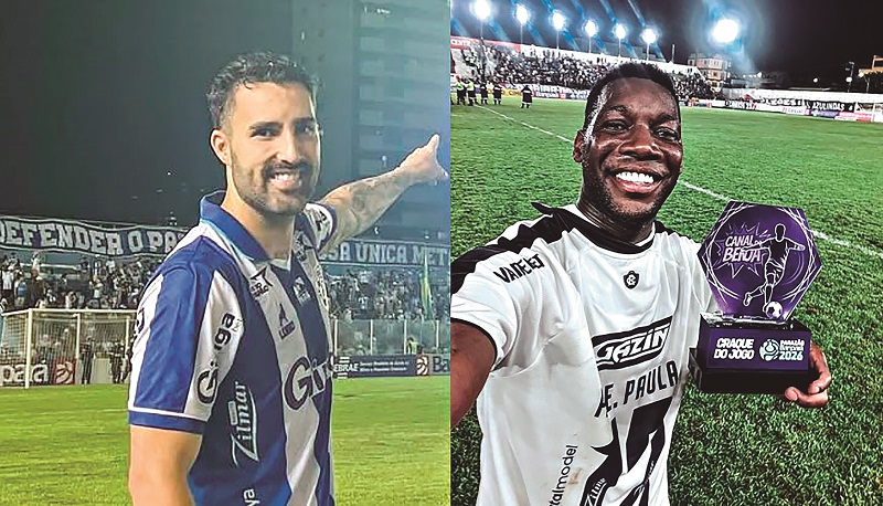 Dois jogadores de futebol sorrindo em campo: um de camisa azul e branca apontando e outro de camisa branca segurando um troféu hexagonal roxo.