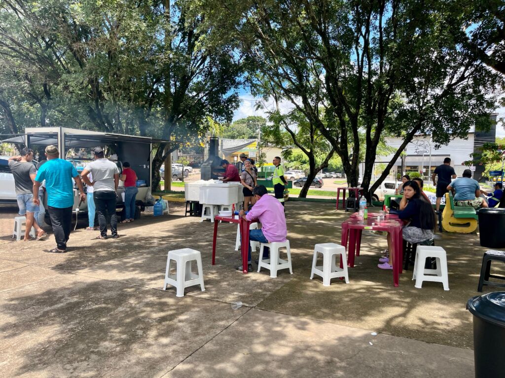 Pessoas em um evento ao ar livre com food truck e mesas sob árvores.