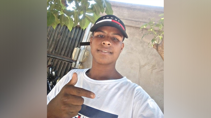 Jovem com boné preto e camiseta branca apontando para a frente.