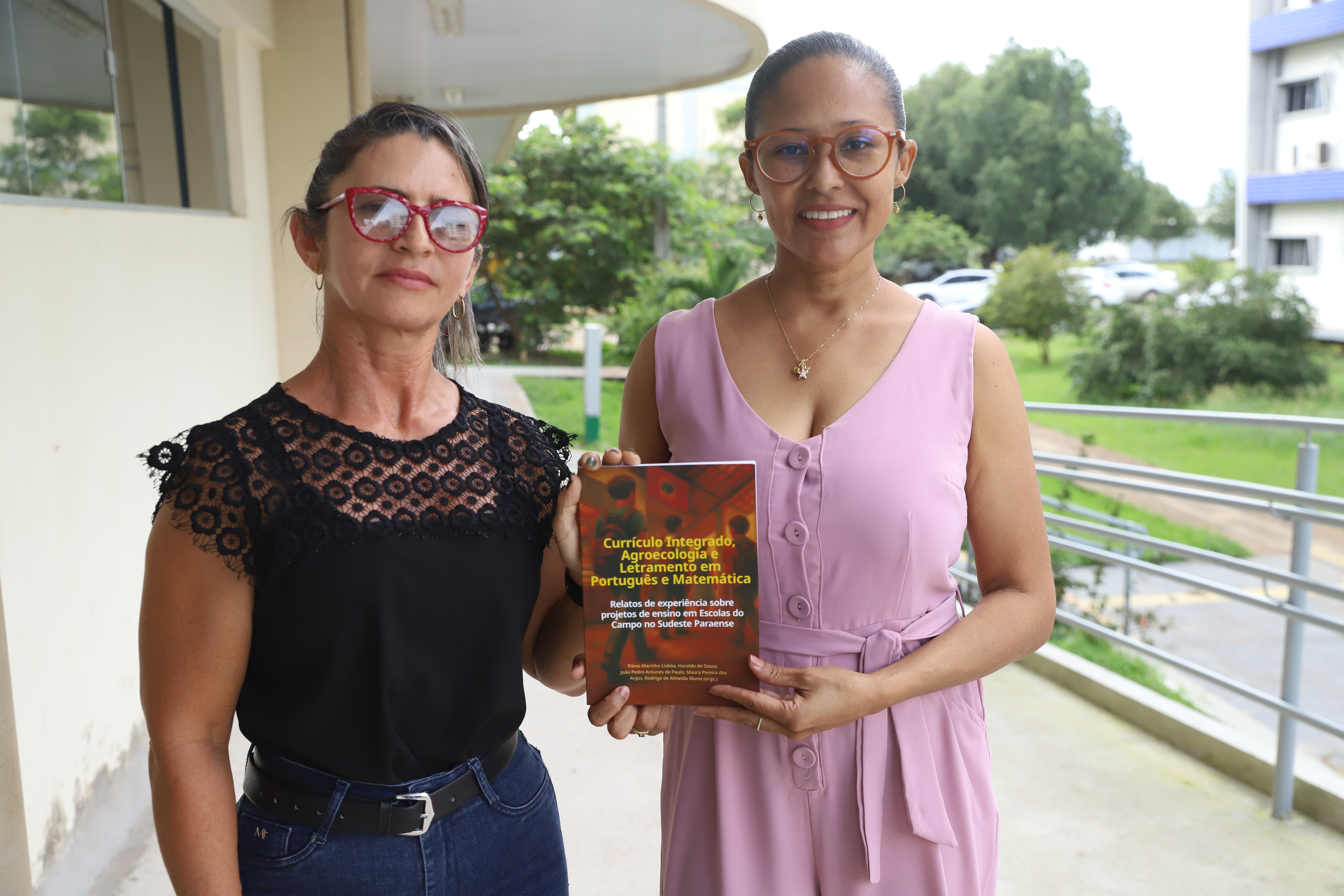 Duas mulheres sorrindo, segurando um livro sobre currículo integrado e agroecologia, em um ambiente externo.