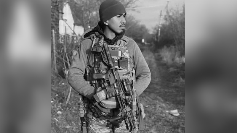 Homem jovem com equipamento militar e rifle em ambiente externo, preto e branco.