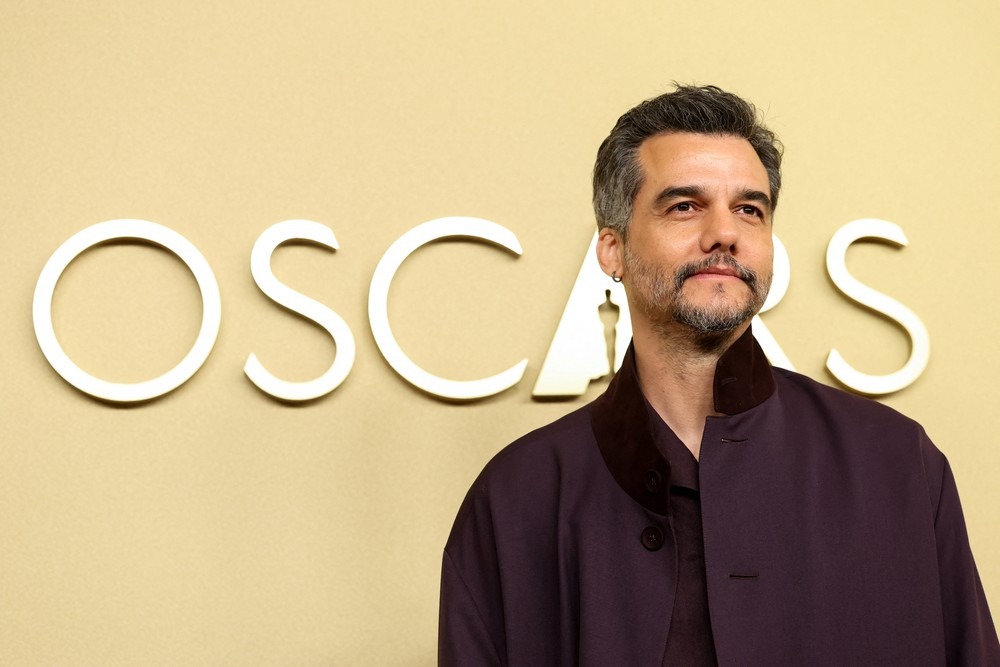 Wagner Moura com casaco roxo e barba, em frente ao logo do Oscar.