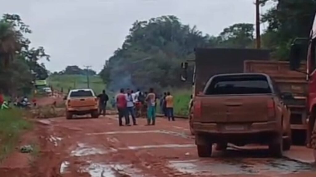 Grupo de pessoas e duas picapes em estrada de terra rural com mata ao fundo, sob céu nublado.