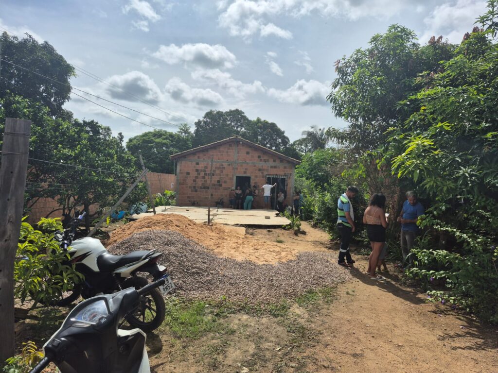 Casa de tijolos com pessoas na entrada e materiais de construção no pátio, em área verde.