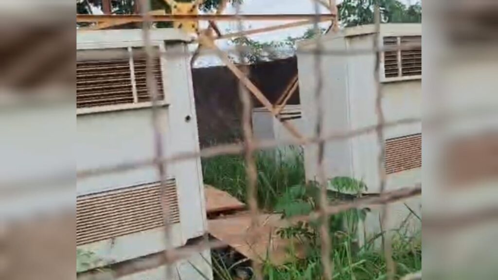 Estrutura modular abandonada e cercada por mato alto, vista através de uma grade borrada.