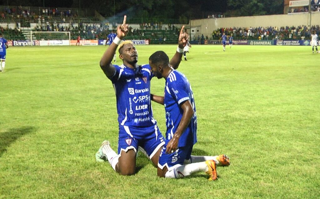 Dois jogadores de futebol em uniformes azuis ajoelhados no campo, um deles celebrando com os braços erguidos.