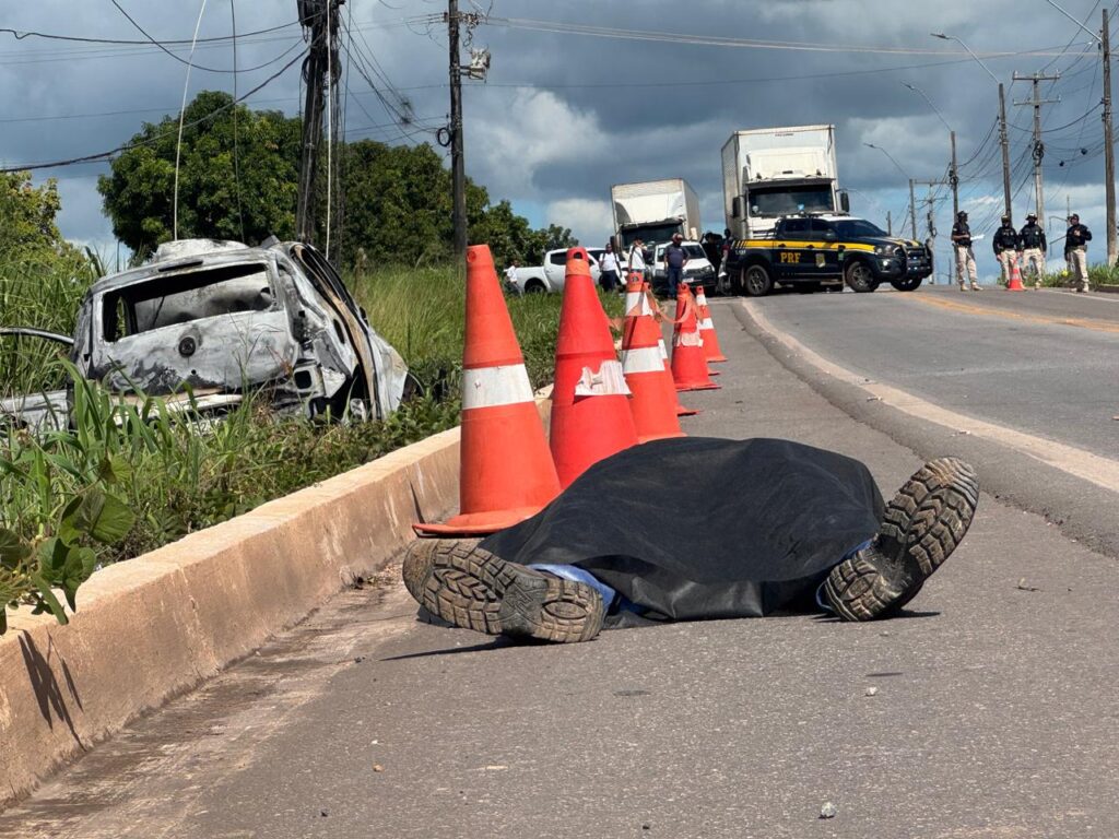 Corpo coberto e carro queimado em cena de acidente na estrada.