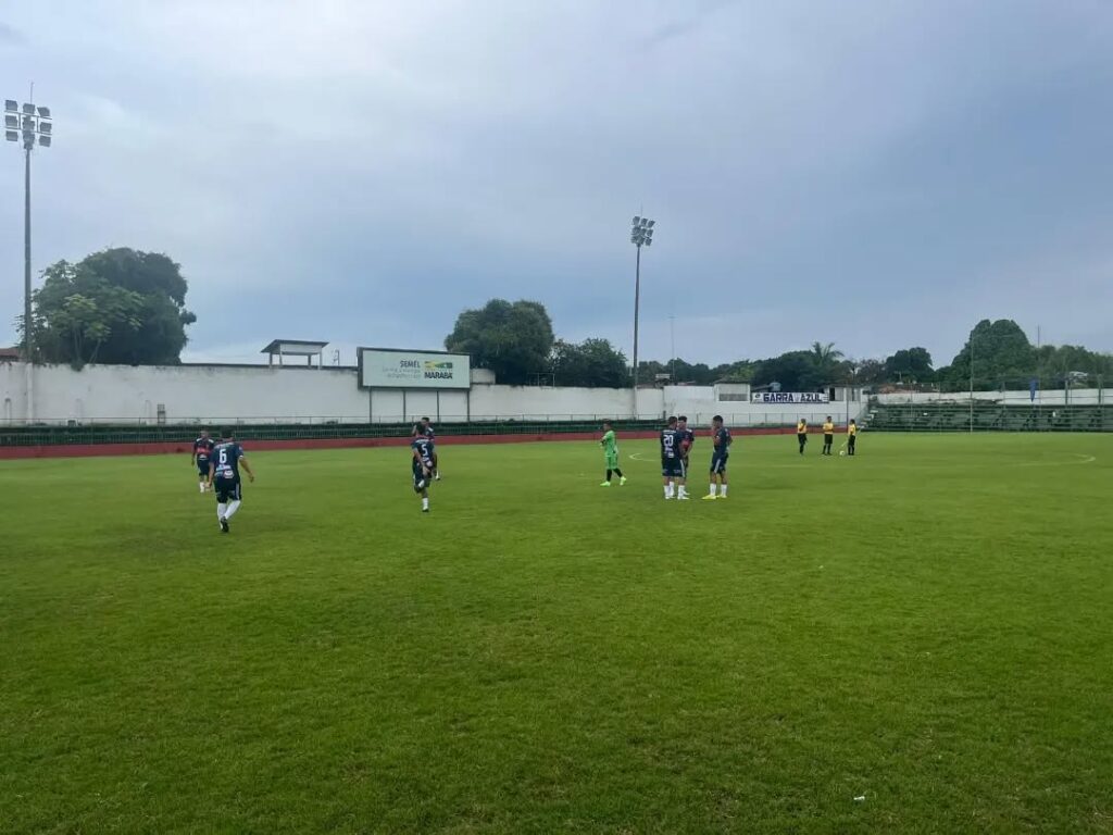 Jogadores de futebol em campo de grama verde com arquibancada e céu nublado ao fundo.