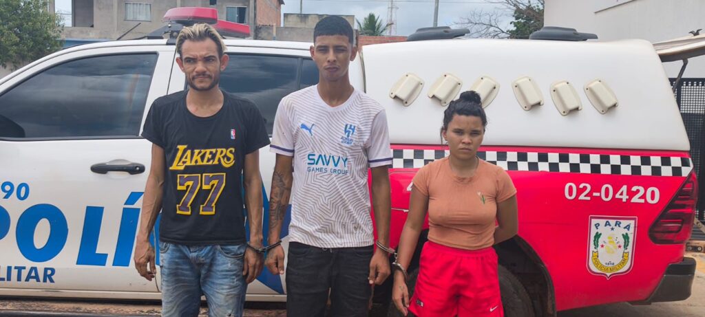 Três pessoas algemadas, dois homens e uma mulher, em frente a uma viatura policial.