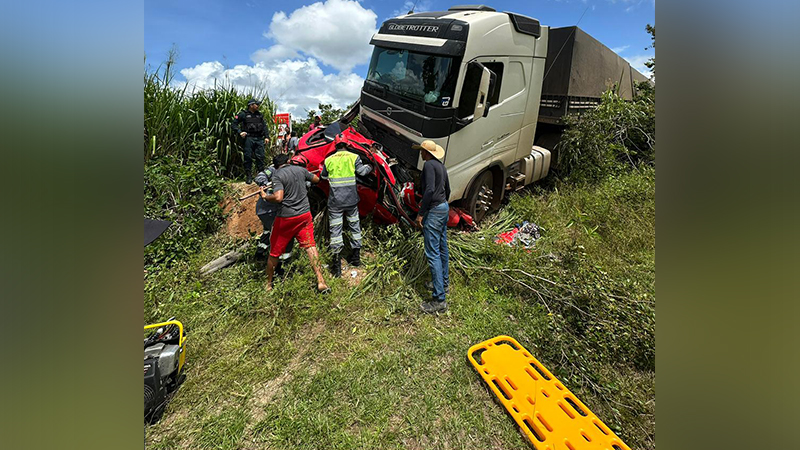 Cena de acidente grave entre caminhão branco e carro vermelho destruído em área rural.