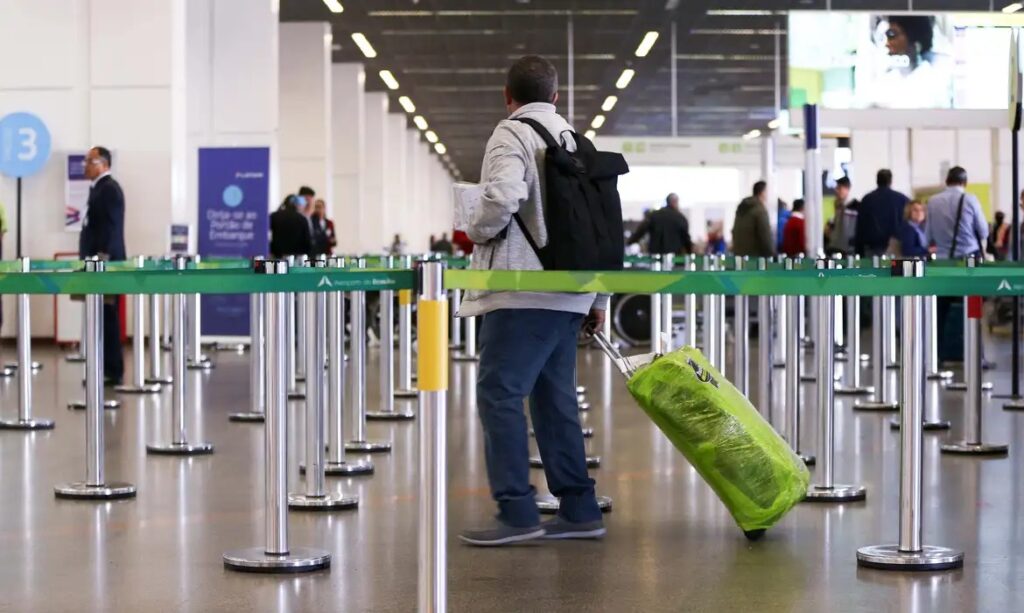 Homem de costas em um aeroporto puxando uma mala verde embalada.