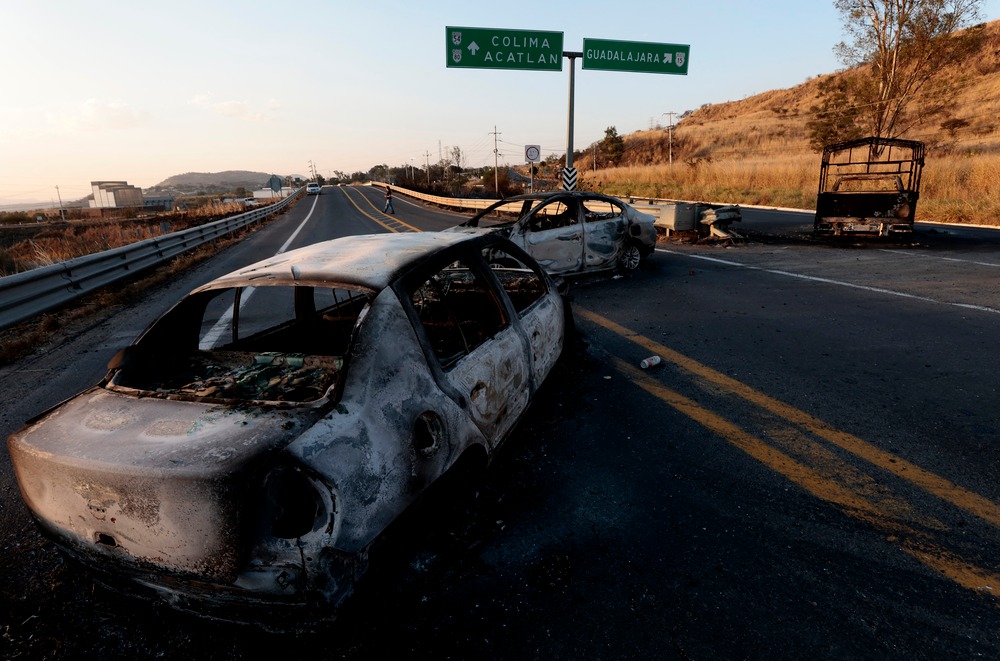 Dois carros queimados e destruídos na beira de uma rodovia, com placas de trânsito indicando Colima, Acatlán e Guadalajara ao fundo.