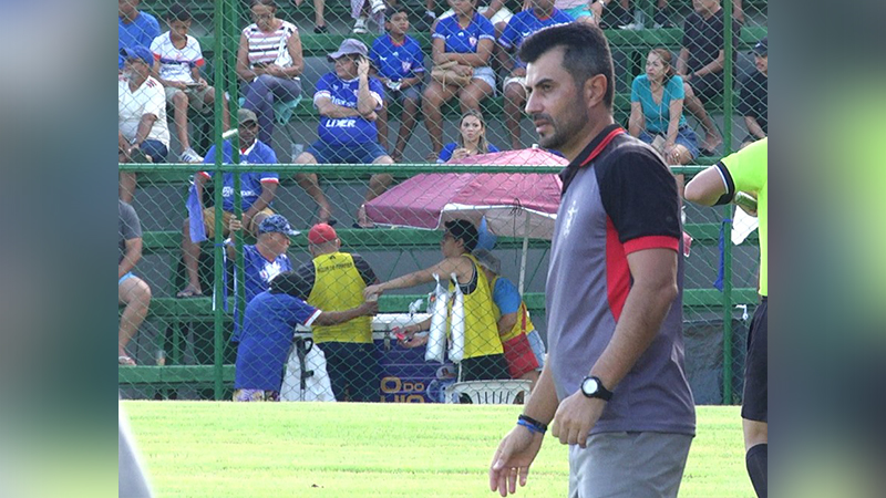 Homem em camisa polo cinza e preta em um campo de futebol, com torcedores ao fundo.