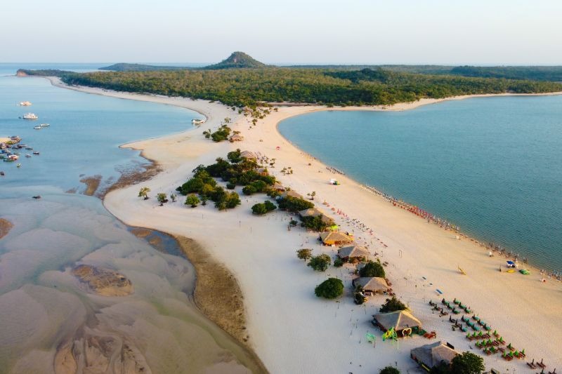 Vista aérea de uma praia fluvial com faixa de areia branca e águas azuis, com cabanas e vegetação.