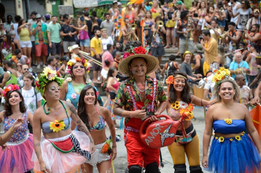 Pessoas em fantasias coloridas e coroas de flores celebram o Carnaval em um bloco de rua movimentado. Um homem sorridente com chapéu de palha e macacão vermelho segura um objeto com 'AMOR' escrito.