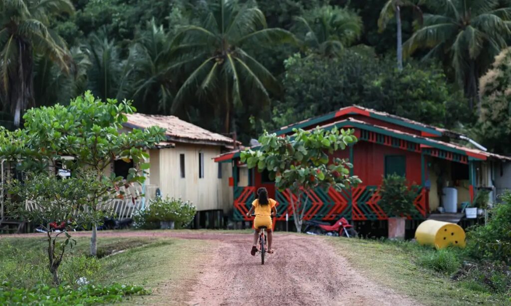 Menina de amarelo pedalando em estrada de terra com casas coloridas e vegetação tropical ao fundo.