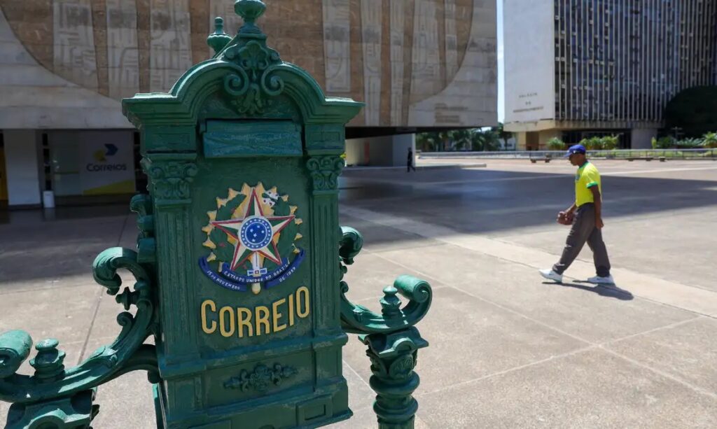 Caixa de correio verde dos Correios com Brasão do Brasil em praça, com edifício governamental ao fundo.