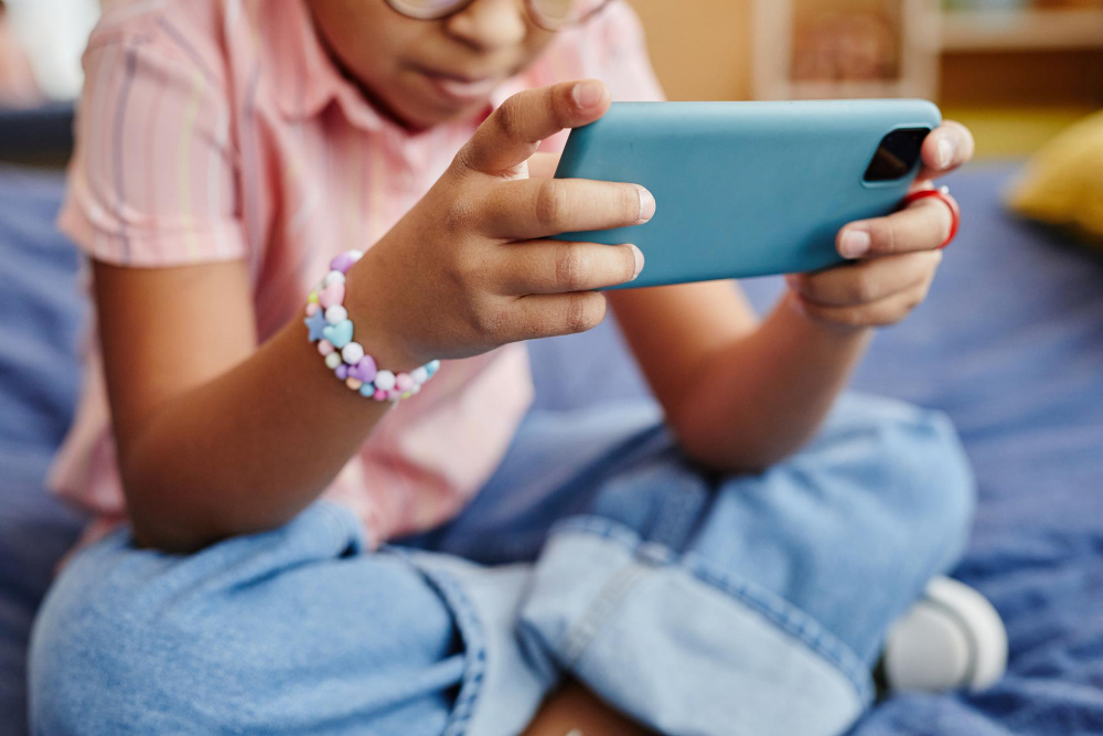 Mãos de uma criança segurando um smartphone azul-claro na horizontal, com um bracelete colorido no pulso.