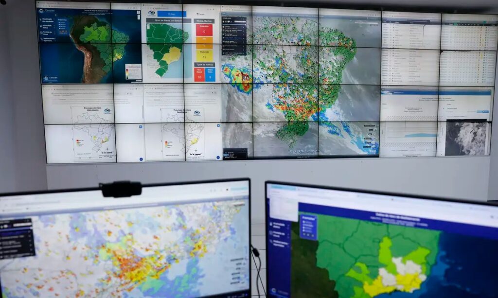 Sala de monitoramento com painel de vídeo exibindo mapas climáticos e dados do Brasil.