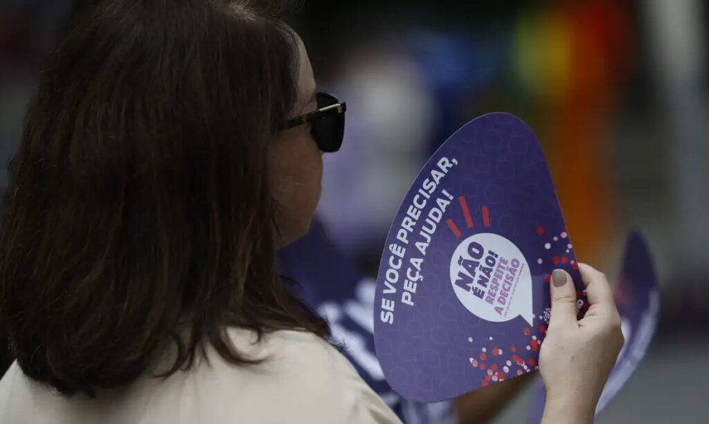 Mulher segurando leque roxo com as frases 'NÃO É NÃO! RESPEITE A DECISÃO' e 'SE VOCÊ PRECISAR, PEÇA AJUDA!'