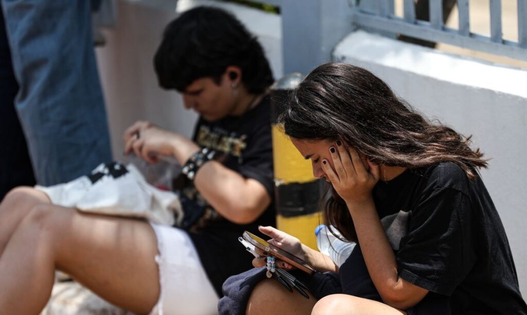 Duas pessoas sentadas ao ar livre, uma mulher no primeiro plano olhando para o celular e um homem ao fundo focado em algo nas mãos.