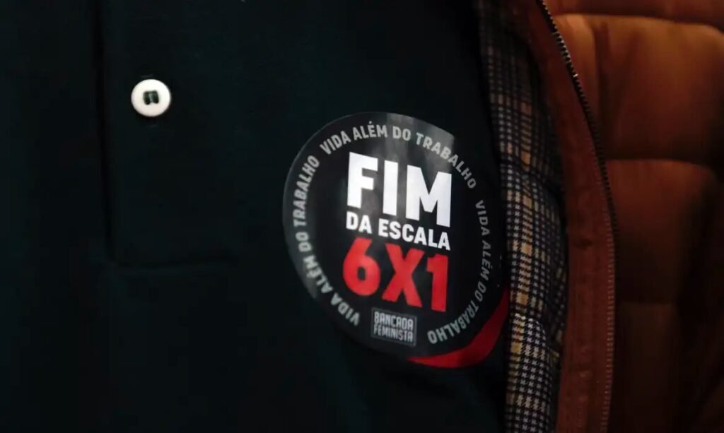 Botton preto com a frase 'FIM DA ESCALA 6X1' em vermelho e branco, e 'VIDA ALÉM DO TRABALHO' em volta, fixado em uma peça de roupa escura.
