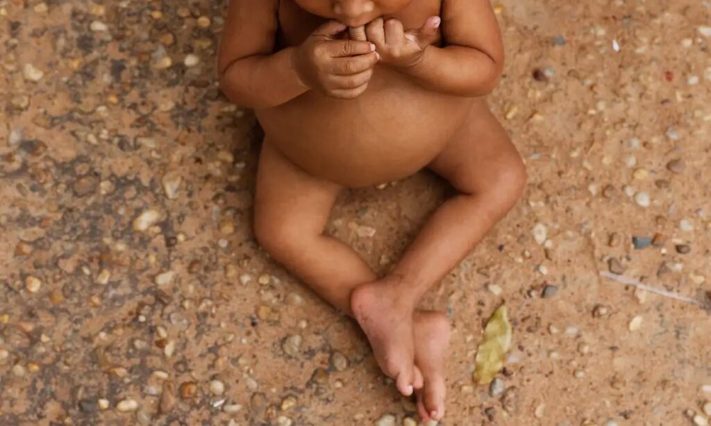Bebê com pele morena deitado em chão de terra com pedras e uma folha, com as mãos na boca e as pernas cruzadas.