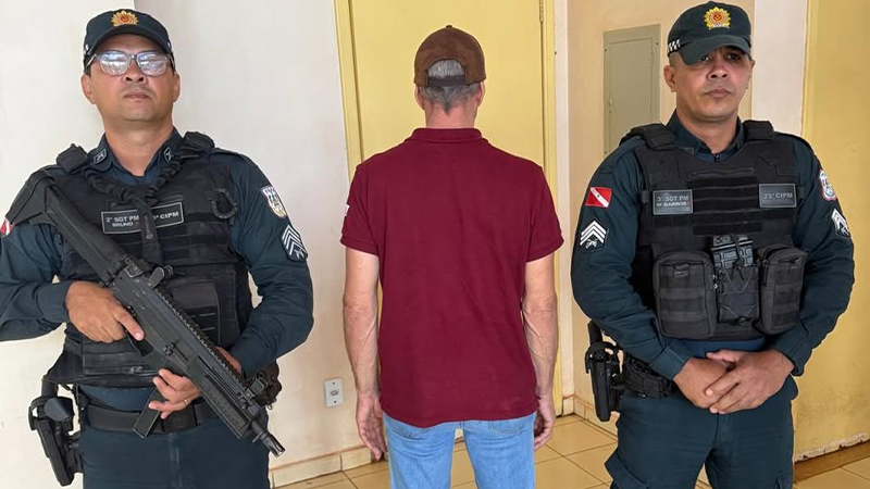 Dois policiais militares com coletes táticos flanqueiam um homem de costas, que usa camisa polo vinho e boné.