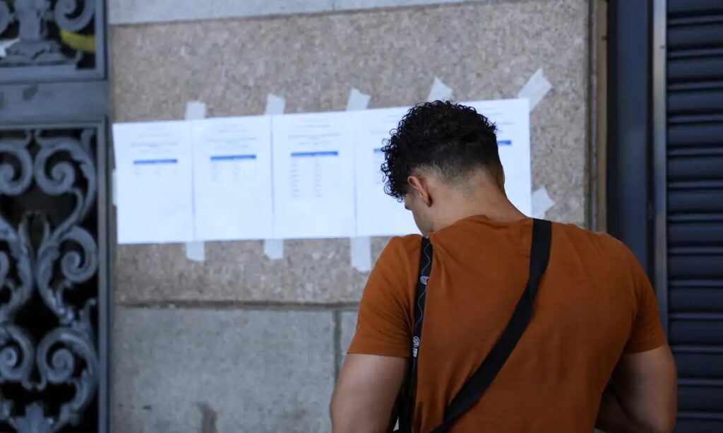 Jovem de costas examinando resultados afixados na parede.