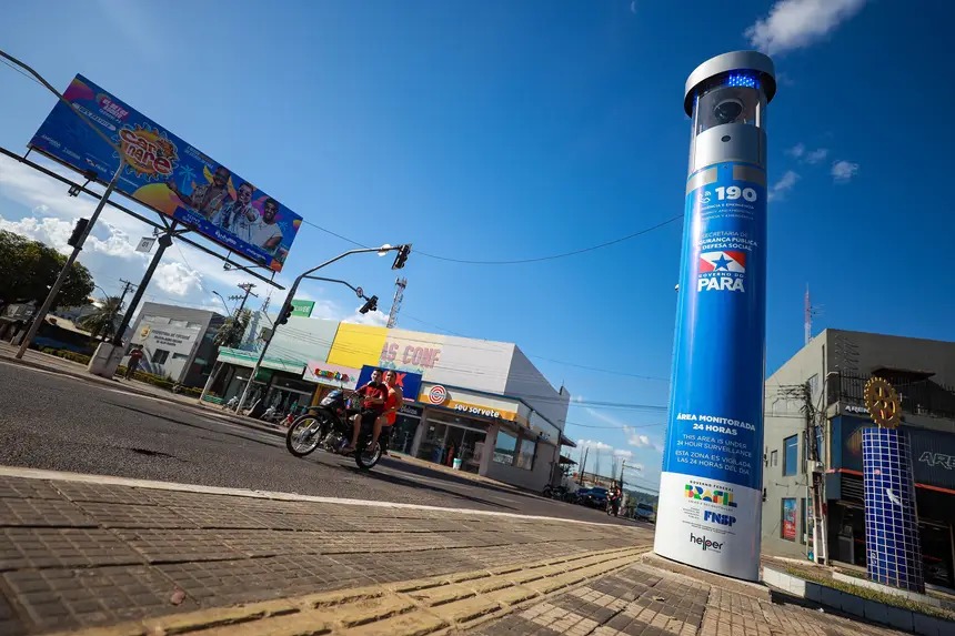 Poste azul de segurança com câmera e informações sobre monitoramento 24h em rua urbana com moto e letreiros.