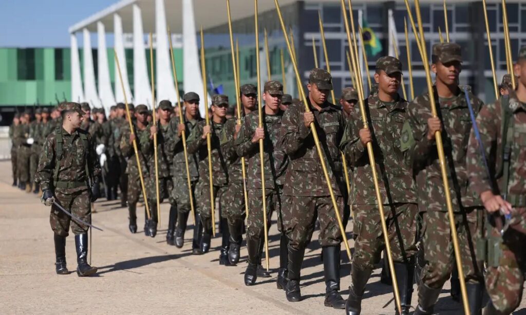 Soldados do Exército Brasileiro em uniforme camuflado marchando com lanças cerimoniais em frente a um prédio governamental e a bandeira do Brasil.
