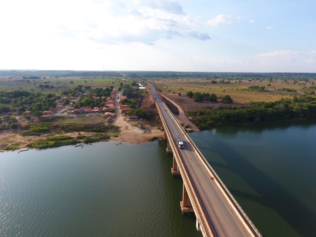 Ponte sobre rio, com caminhão, vilarejo e paisagem rural sob céu azul.