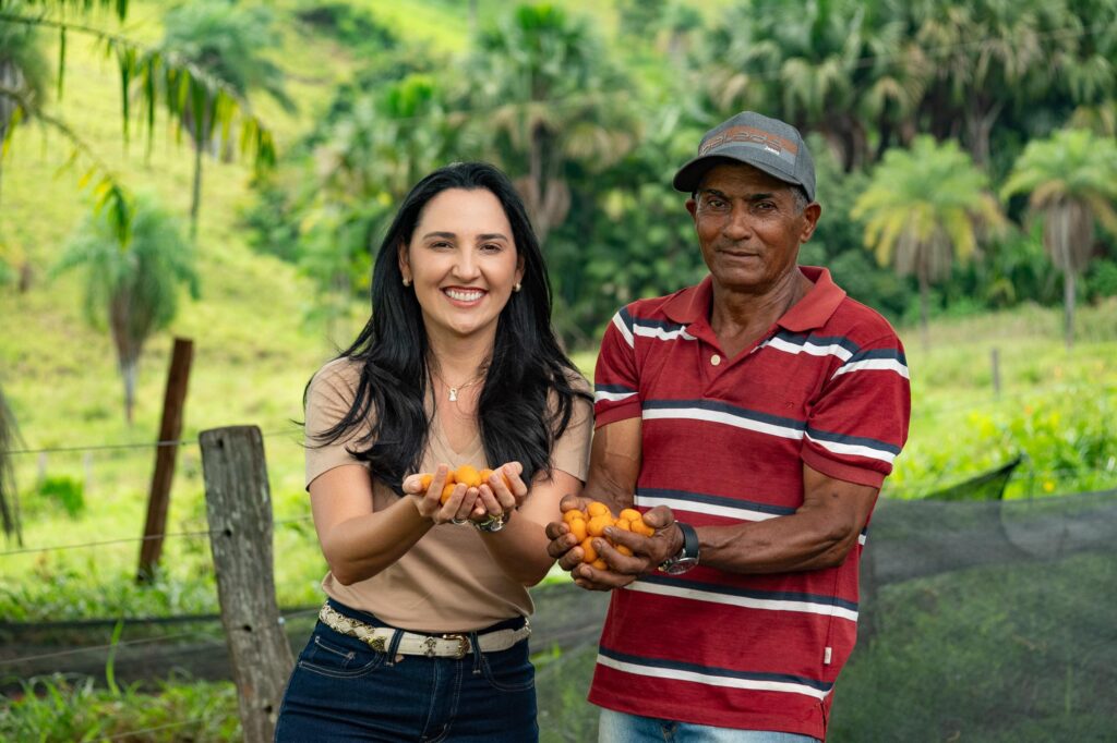 Mulher e homem sorridentes segurando frutas alaranjadas em campo verde.