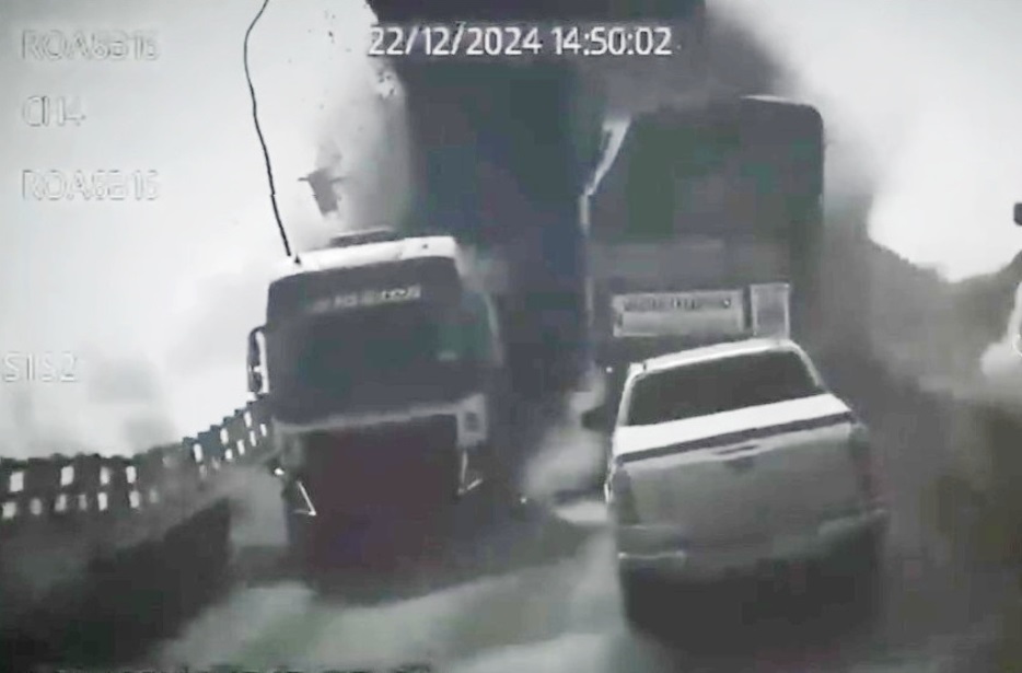 Registro de câmera de segurança mostrando colisão ou incidente envolvendo caminhões e uma picape em uma ponte, com fumaça.